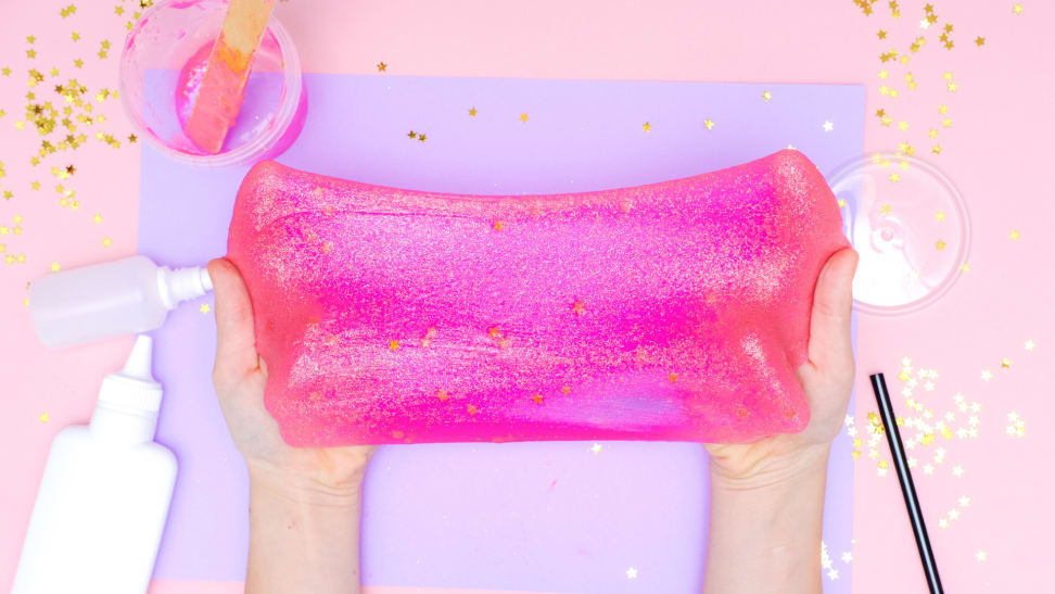 Hands stretching pink glitter slime with glue, mixing materials, and glitter in the background.