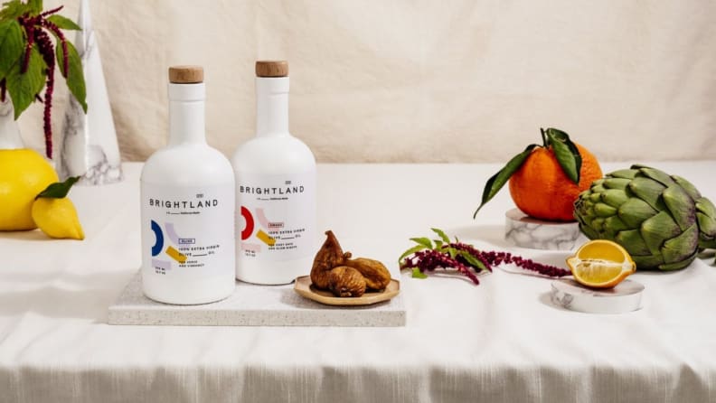 On a table, two bottles of Brightland olive oil are on display, alongside some fruits and vegetables such as an artichoke, lemon, and oranges.