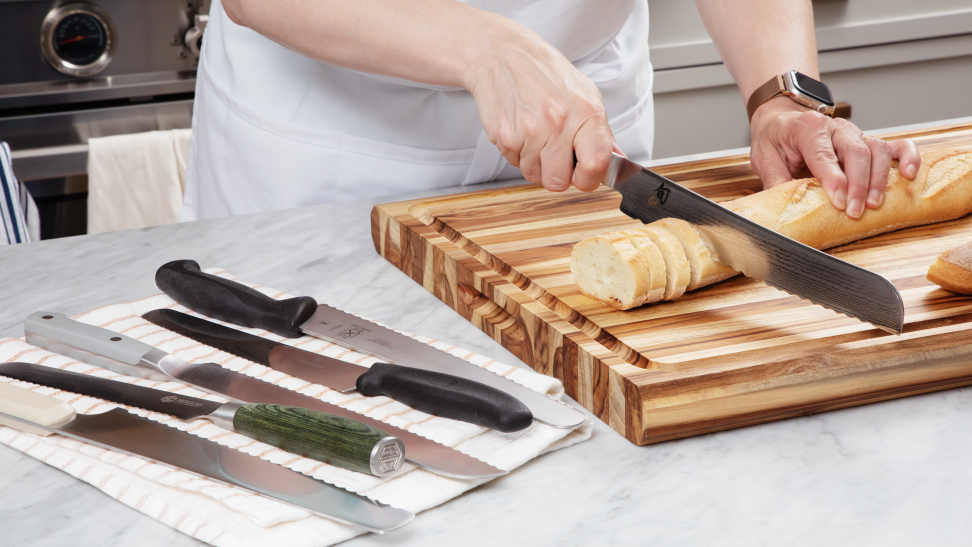 Hand cutting a baguette with a serrated bread knife, beside several other bread knives