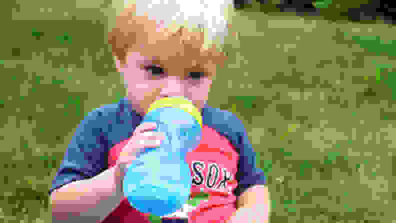 A blonde child in a Red Sox shirt sits on grass and sips through a blue Nuby Clik-it Flex Straw cup.