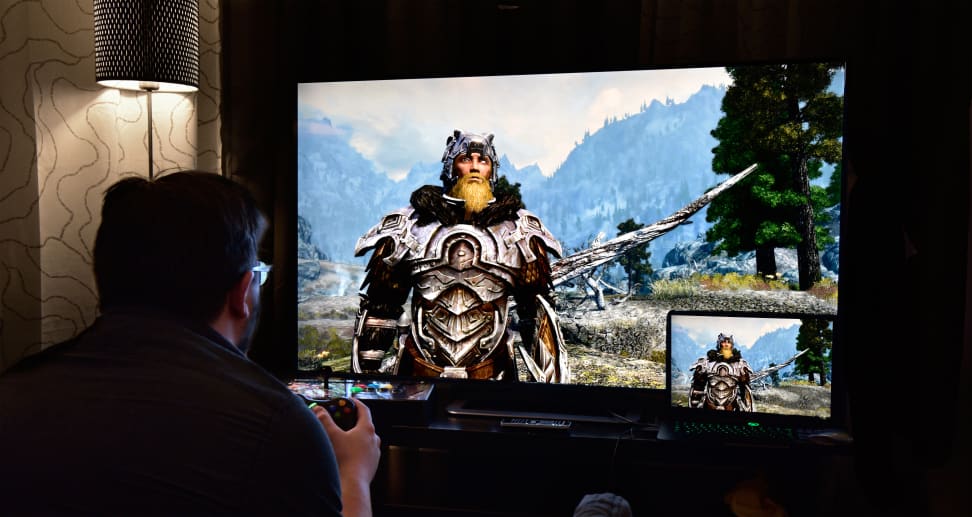 A man sits in a dark room playing a video game with a Viking-like character on the screen
