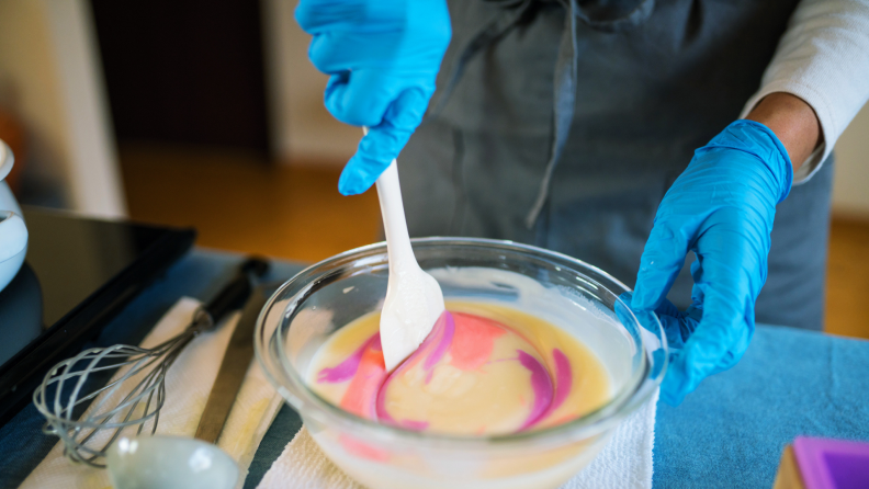 Person wearing gloves using spatula to stir together homemade soap in glass bowl