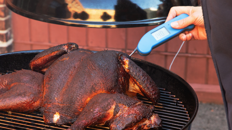Person using a Thermapen to check the temperature of smoking turkey on a charcoal grill outdoors.
