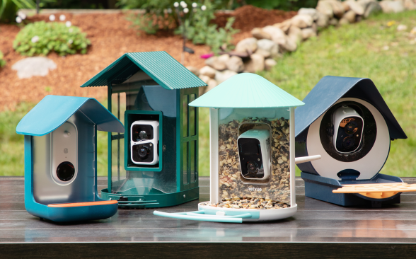 Four bird feeders in blue and green shades sit in a group on a brown table with a garden and green grass in the background