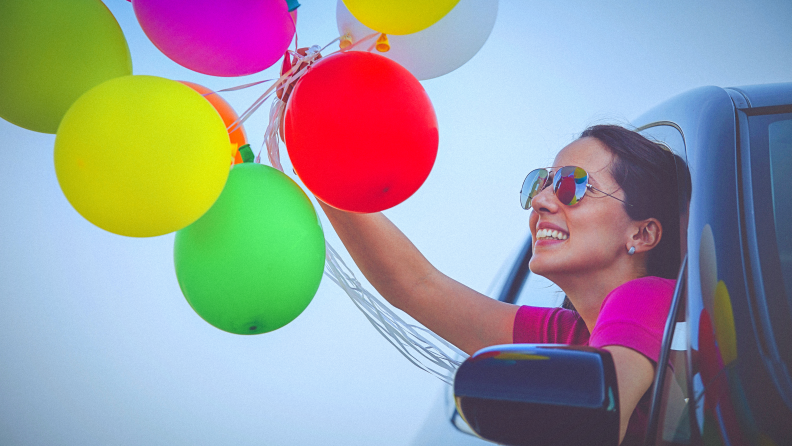 A woman smiles as she hangs out of a car window with colorful balloons