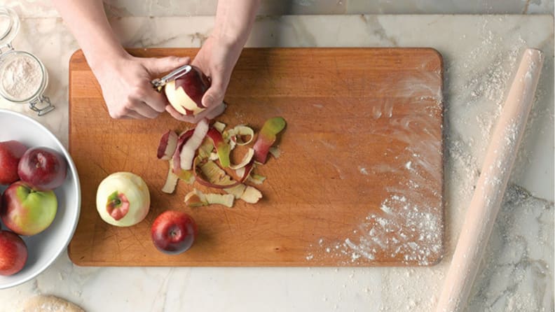 Two hand peeling an apple on a cutting board, with a rolling pin closeby