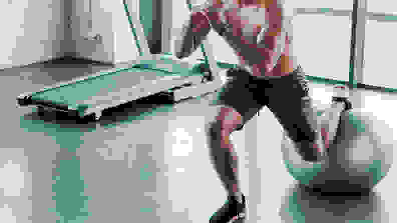 A muscular athlete does lunges with one leg balanced on a medicine ball. A treadmill is nearby in the background.