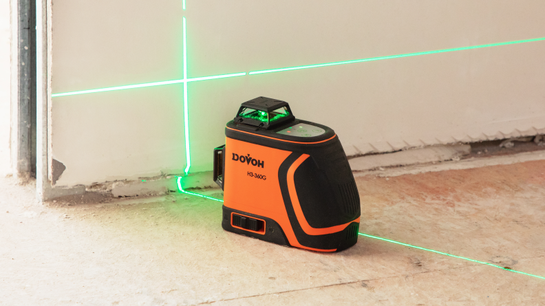 An orange laser level shoots a crossed green laser onto a wall.