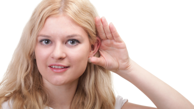 A woman with blond hair holds her hand to hear ear, gesturing that she can't hear