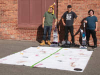 Three people stand in front of a brick wall and next to a white rug with stains on it.