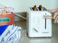 A Cuisinart 2-slice toaster is being shown in close-up. A hand demonstrates the toaster's operation; two slices of toast are ready to be retrieved from the slots.