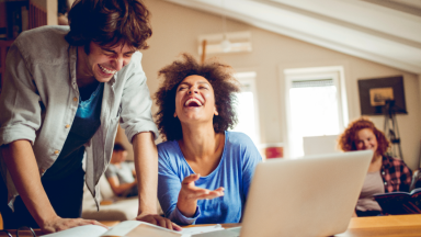 Two friends laughing together while working on a laptop at home, highlighting the fun and connectivity possible with T Mobile 5G Home Internet.