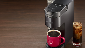 在棕色背景下，有一台黑色不锈钢Keurig单杯咖啡机正在煮咖啡。在机器旁边，有一杯冰咖啡。