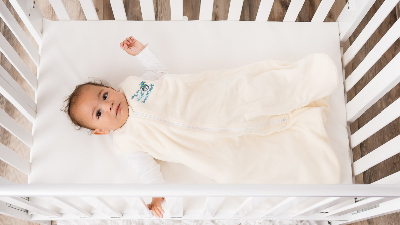 A baby wearing a cream onesie lays in a crib.