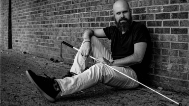 'The Blind Life' host Sam Seavey shown sitting up against a brick wall holding a guide cane.