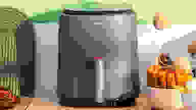 A Cosori Lite four-quart air fryer sitting on a kitchen counter in front of a green wall with white tiles and surrounded by food at home.