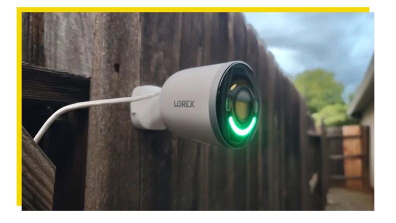Lorex 4K Spotlight mounted on wooden fence outdoors with green LED light.
