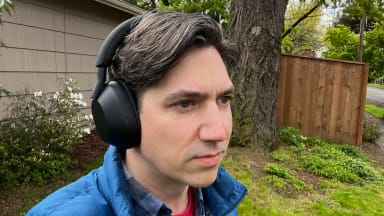 A man with brown and grey hair wears the matte black Sony WH-1000XM5 headphones in front of a large tree and a beige house.