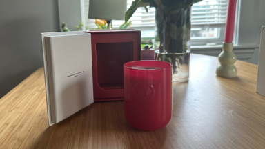 A Glossier You candle in a red glass jar sitting on a table in front of its box.