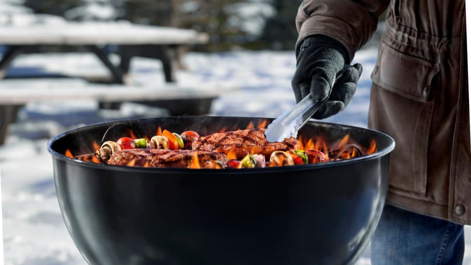 Outdoor grill in the wintertime with gloved hand using tongs to flip meat and vegetable skewers