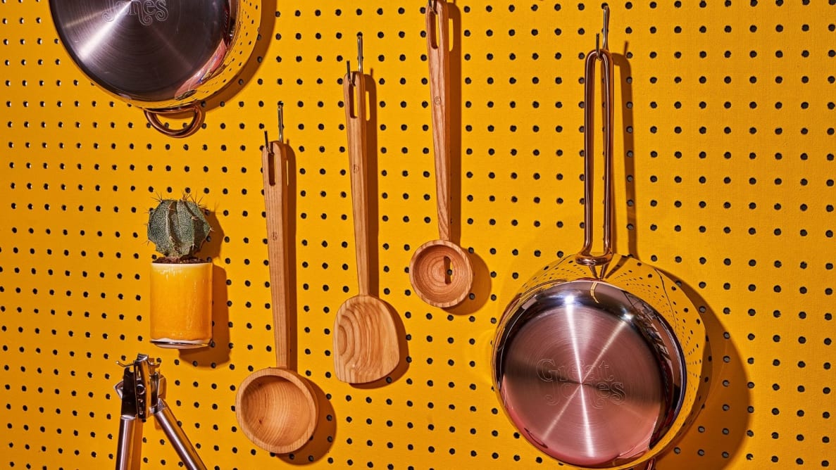 Wooden kitchen utensils hang next to a stainless steel saucepan on a bright yellow pegboard.