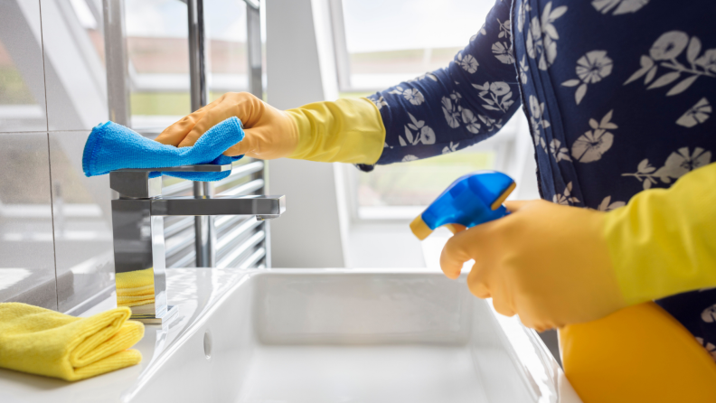 Person with yellow rubber gloves cleaning bathroom sink with a spray nozzle and microfiber cloth
