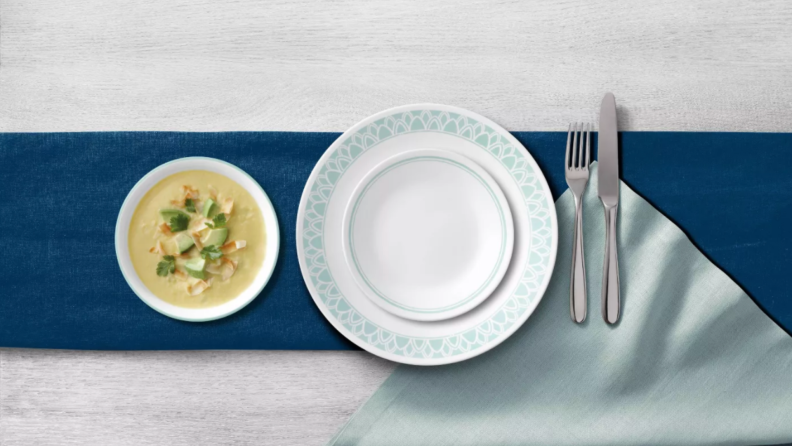 Two empty white and aqua colored plates stacked together next to bowl of soup and silverware.