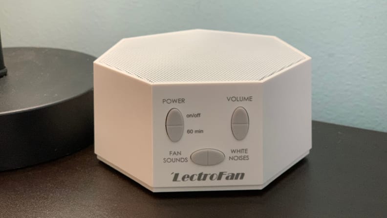 A white hexagon Adaptive Sound LectroFan sound machine with top speakers and buttons on the front sits on a night stand.