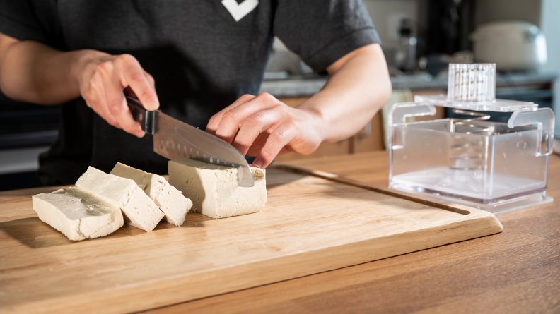 A person cutting a block of tofu on a cutting board with a chef's knife.
