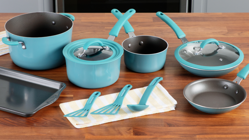 Turquoise blue colored pans, pots, lids and cooking utensils next to small rectangular baking sheet on top of wooden countertop surface.