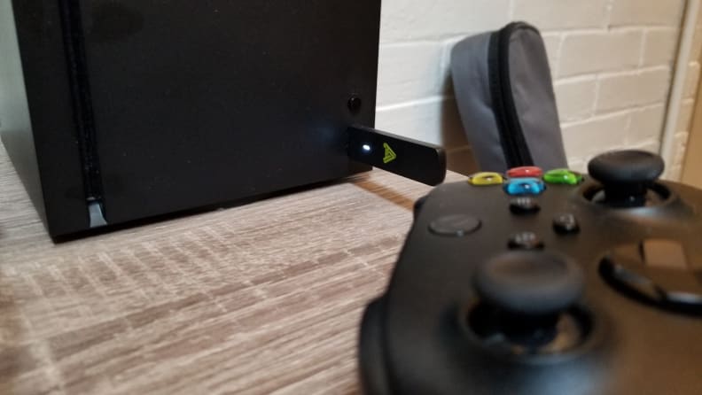 A USB dongle is inserted into the front of an Xbox Series X console. In the foreground, out of focus, is an Xbox controller.