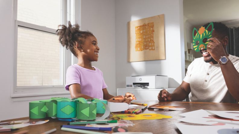 A young girl and her dad laugh while making crafts at a table, with an HP printer in the background.