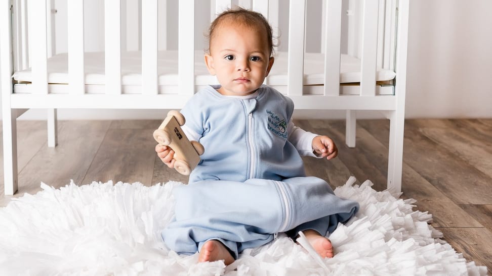 An infant wearing a blue Magic Merlin's Magic Dream Sack sits in front of a crib.