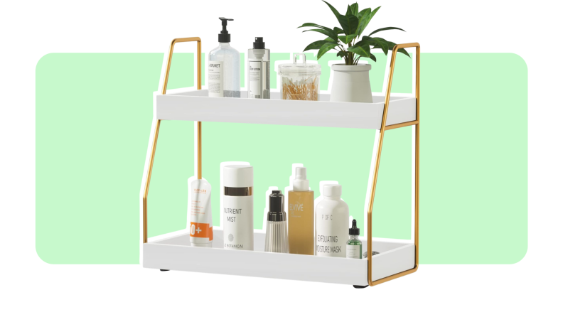 A white and gold two-shelf unit with beauty products on top in front of light green background..