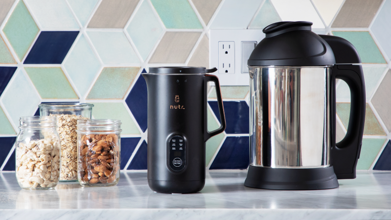 The Nutr plant-based milk maker next to the Almond Cow plant-based milk maker on granite countertop in front of tiled wall and 3 clear jars with snacks inside.