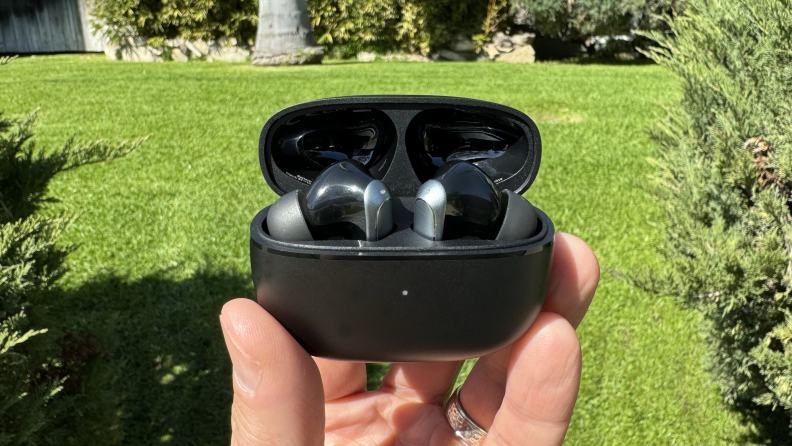 A hand holding the EarFun Air 2 earbuds in their case outside in the sunlight with green grass and bushes in the background.