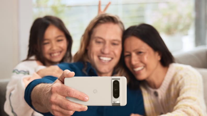 A man, woman and child sit on a couch in their home. The man, sitting between the two females, is holding his arm out, with a Google Pixel 8 Pro in his hand. The three people are smiling for a selfie.