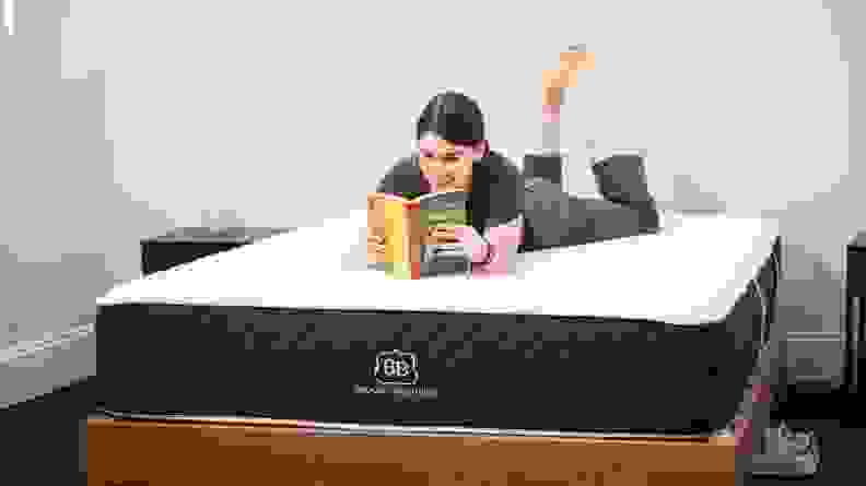 A woman lying on her stomach on a bare mattress, reading a book.