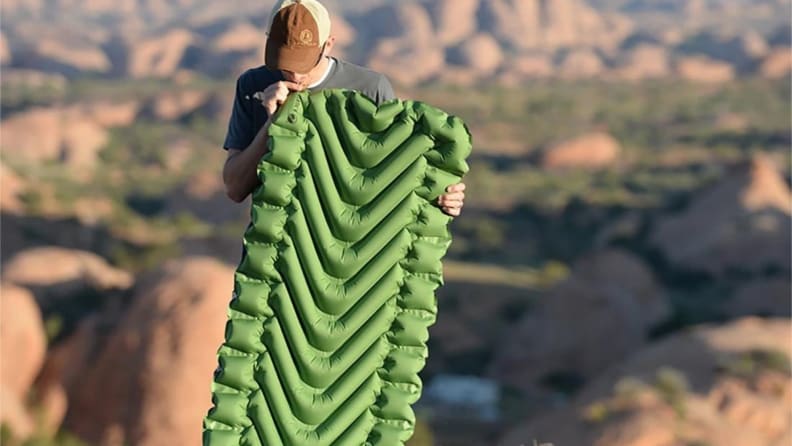 A man blows up a green sleep pad on a mountainside.