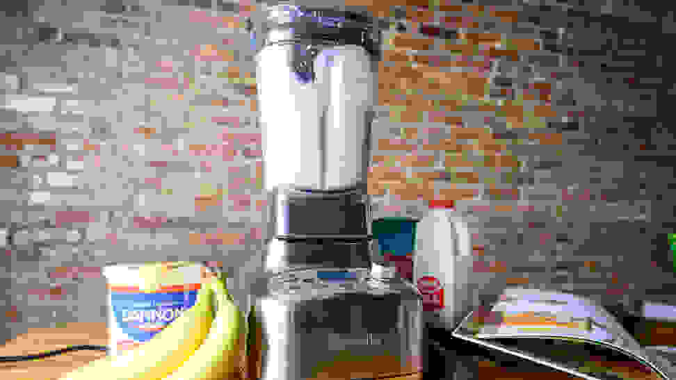 A strawberry banana smoothie is being made in the Breville Super Q blender with a container or strawberry yogurt, bananas, and cookbook next to it.