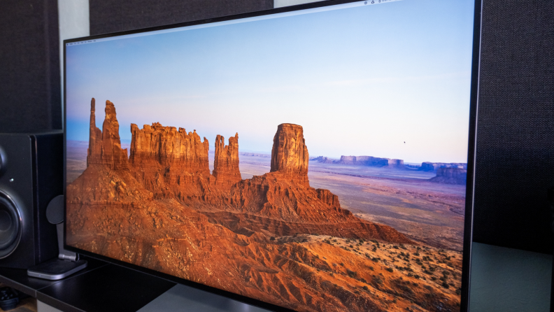 A close-up angled view of a widescreen monitor displaying red rock formations in a desert, with part of a black studio speaker visible beside it.