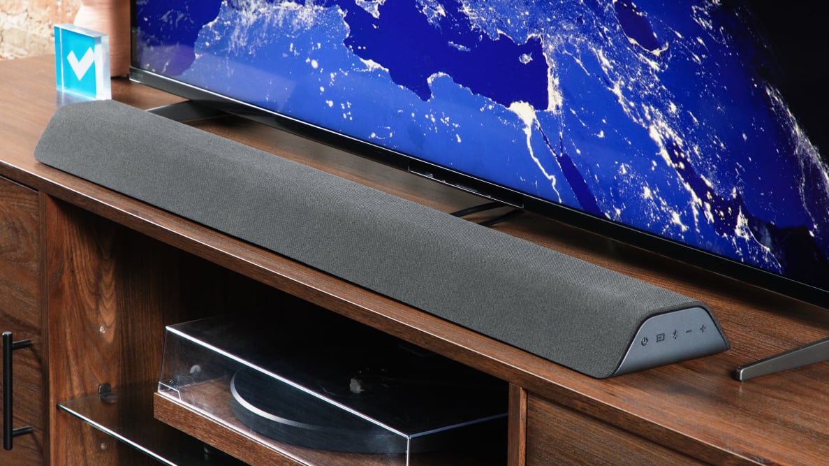 Angled shot of the Vizio M-Series All-in-One M213AD-K8 Dolby Atmos Soundbar on an entertainment center in front of a TV.