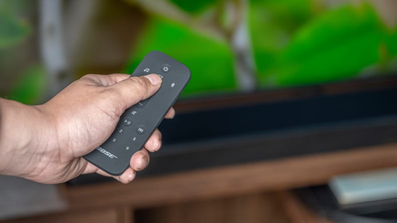 A hand holds a rubberized remote above an all black soundbar on a wooden console set beneath a TV