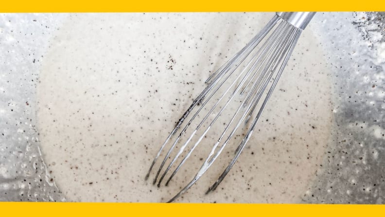 A whisk leans against the side of a bowl filled with waffle batter.