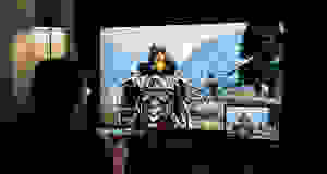 A man sits in a dark room playing a video game with a Viking-like character on the screen