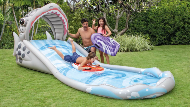 Father playing with children on the Surf ‘N’ Slide Shark Water Slide while son uses inflatable toy to slide.