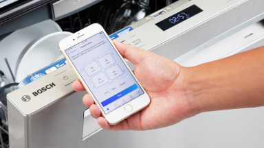 A Bosch dishwasher and a hand holding a smartphone displaying the Bosch app.