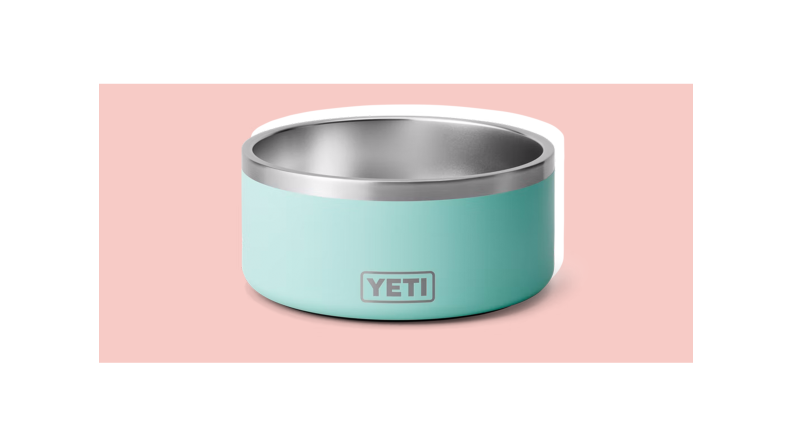 A metal dog bowl with a light blue casing and the name "Yeti" against a pink background.