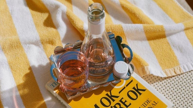 A small glass of wine is sitting next to a small wine bottle atop a white and yellow striped towel with a book nestled underneath. It's a very sunny, almost beach-like scene.