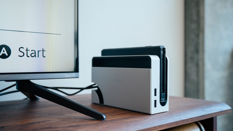 A nintendo switch oled in a dock next to a tv on a credenza